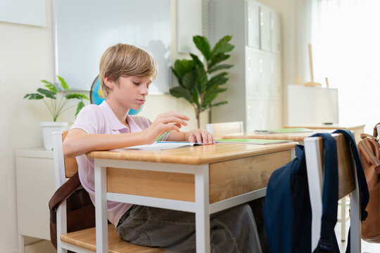 Caucasian schoolboy doing homework in primary classroom, Young boy focused on writing in notebook at school desk, Dedicated student studying and writing assignment in classroom