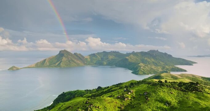 Aerial drone view of Fiji's ocean bay, lush green islands and rugged volcanic peaks framing a tranquil turquoise eater beneath a vibrant rainbow arching across the tropical sky. Tropical paradise