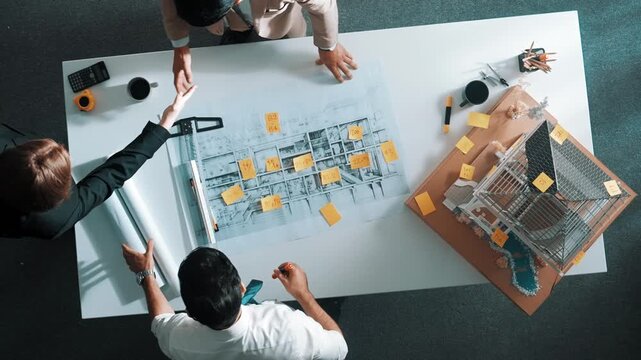 Top down view of architect engineer writing building structure idea on note and stick on blueprint. Aerial view of manager working on notes while consulting team about house structure. Alimentation.