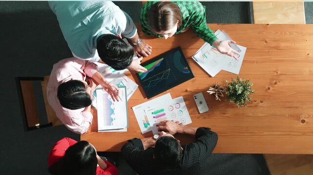 Top view of skilled business man standing while looking financial chart document at meeting while start up team with casual cloth discussing and analyzing investment statistic at table. Convocation.