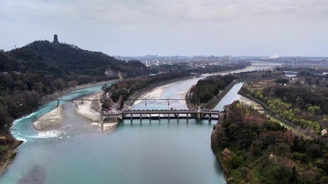 A river diversion hydraulic structure splits water into channels, with a forested hill and distant town in the landscape.