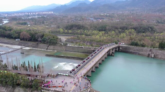 A historic hydraulic project site with a stone bridge spanning a turquoise river, surrounded by mountains and vegetation, with visitors on the bridge and square.