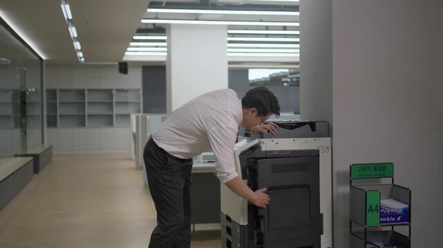 A businessman in a white shirt leaning over a printer machine to collect documents in a modern office while working overtime at night.