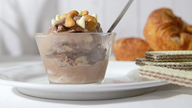 una copa de cristal con Helado de chocolate con chispas de chocolate blanc, almendras y nueces de la india encima, con panecitos y pared de madera en el fondo, una cuchara dentro de la nieve.
