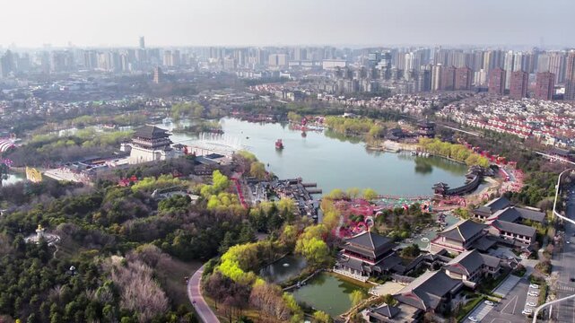 This aerial shot shows a large traditional garden with a central lake, set against a modern urban city skyline in the distance.