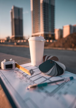 Architectural plans, color palette samples and takeaway coffee placed outdoors near modern residential towers. Concept urban development, project planning, construction management and design workflow.