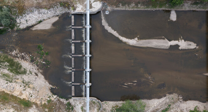 Aerial view of a concrete river dam showing low water levels during a summer drought, with exposed sandy riverbanks and sparse vegetation in Portugal.