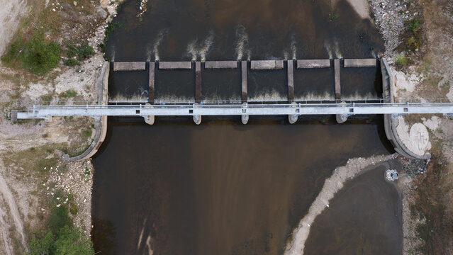 Aerial view of a river dam with low water levels during a summer drought, showing concrete structures and dry riverbanks in Portugal.
