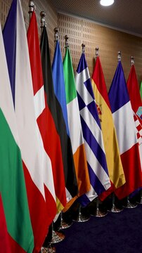 A group of flags from different countries stands in a conference room. Each flag represents a nation and is arranged neatly. The lighting adds a formal touch to the setting.