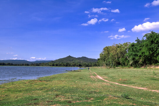 A man walking on a small green path alongside Huai Phueng reservoir under a bright blue sky in Kalasin, Thailand.