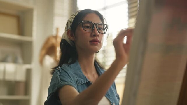 Young Asian woman artist sitting on stool painting blank canvas on wooden easel with palette and brush in stylish home art studio, full body creative lifestyle and hobby concept	