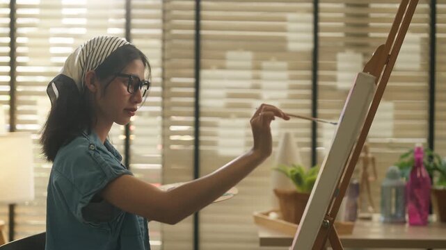 Young Asian woman artist sitting on stool painting blank canvas on wooden easel with palette and brush in stylish home art studio, full body creative lifestyle and hobby concept	