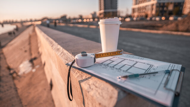 Takeaway coffee, measuring tape and architectural documents capture productive morning routine of construction professionals. Modern buildings symbolize growth, teamwork and successful projects.