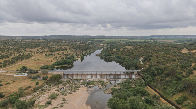 Aerial view of the Acude do Furadouro dam spanning the river, surrounded by lush cork oak forests and dry fields under a cloudy sky in Mora, Distrito de Evora, Portugal.