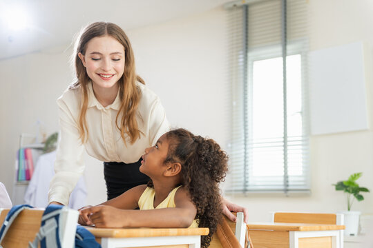 Supportive teacher mentoring children during class lesson, Teacher walking around classroom and helping students with study, Diverse primary school students learning in modern classroom
