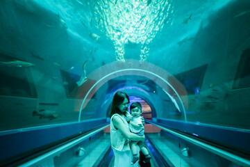 Asian mother holding her baby boy, watching big fish swimming in a giant aquarium tank. © Hide_Studio