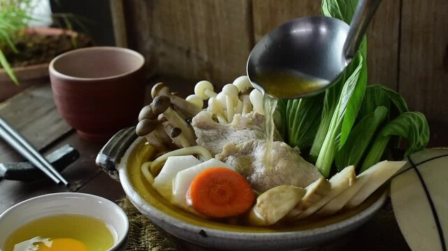Chef pouring hot savory broth from a ladle into a traditional Japanese hot pot filled with fresh pork slices, bok choy, shimeji mushrooms, carrots, and radishes in a rustic clay pot.