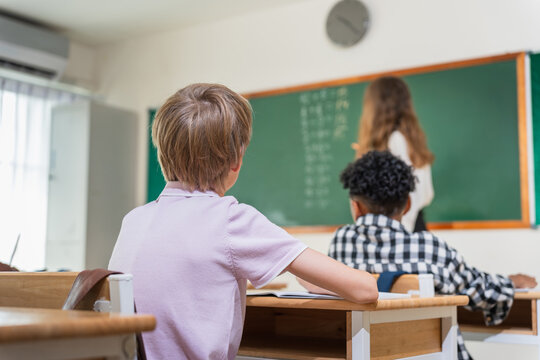 Students in a classroom during a lesson, Teacher conducting a math lesson in a diverse elementary classroom, Primary school teacher writing on chalkboard with diverse pupils in class