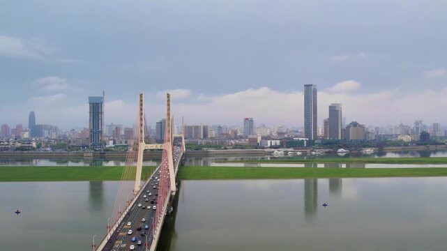 A cable-stayed bridge spans a wide river, with urban high-rise buildings lining the opposite bank and traffic on the bridge. Greenery surrounds the approach ramp.