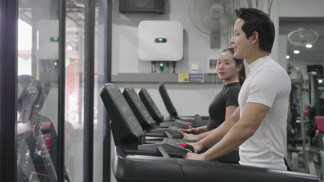 A friendly male trainer is setting up the treadmill display for an Asian woman exercising in a modern gym.