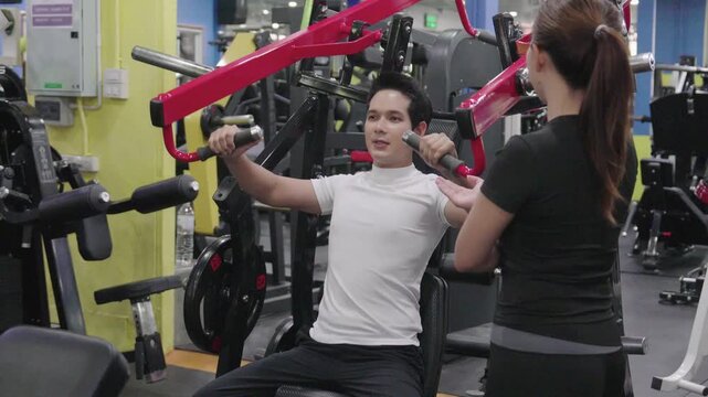 Strong young Asian man lifting barbell on bench press machine with the assistance of a female trainer at the gym.