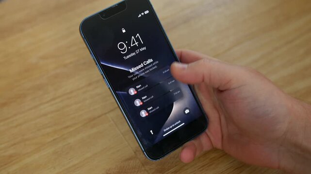 Phone showing missed calls from Nan rests on a wooden table as a hand taps the lock screen. Close-up mobile, cell, smartphone view highlights granny, grandmother, and family contact alerts