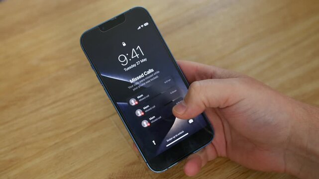 Phone showing missed calls from Mum rests on a wooden table as a hand taps the lock screen. Close-up mobile, cell, smartphone view highlights mother, mom, parent, and family contact alerts