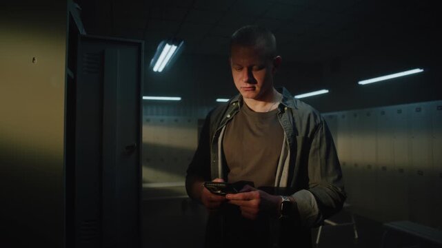 Young Man With Short Haircut Stands In Sunlit Locker Room, Focused On Smartphone Screen, Checking Training Plan Or Music Playlist Before Gym Session. Concept Of Digital Lifestyle In Sports.