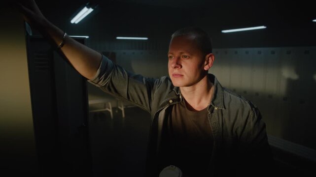 Cinematic Close Up of Focused Male Athlete Leaning Hand on Metal Locker, Looking Thoughtfully Into Window With Warm Morning Light on Face Inside Sports Club. Concept of Authentic Lifestyle Moment.