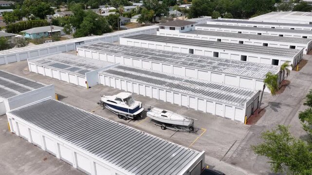 Self-storage complex with white metal unit rows in Bradenton offers outdoor boat storage, solar rooftops, palm trees, and residential neighborhood beyond. Florida, USA. Aerial drone flyover shot