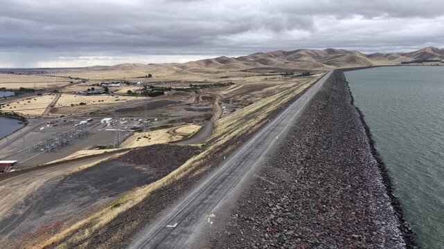 San Luis Reservoir rocky earthen dam stretches toward golden rolling hillsides, calm green water meeting rugged stone embankment near Gustine under heavy storm clouds. Aerial drone wide shot