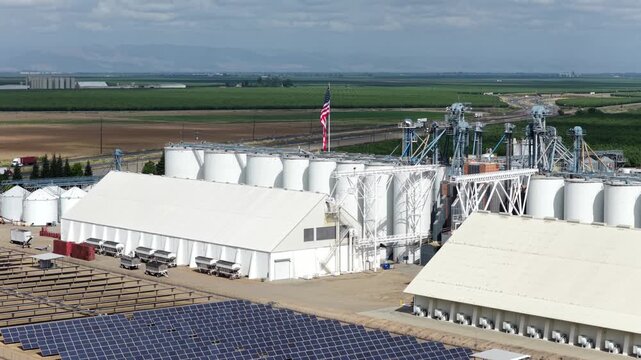 Arbuckle almond processing facility towers above neat solar panel rows, American flag proudly waving beside tall grain silos across flat sunny California farmland scene. Aerial drone rising shot