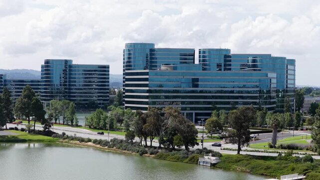 Oracle corporate headquarters towers rise beside calm green lake, modern glass buildings reflecting clouds amid landscaped paths near Redwood Shores California shoreline. Aerial drone wide shot