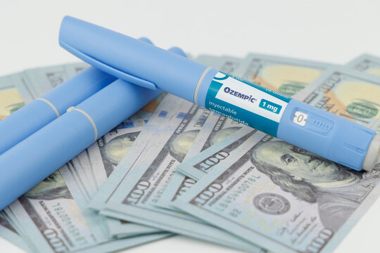 Front angle layout of blue ozempic medication pens and cash dollars for expensive medical health treatments on white background area. In Barcelona -Spain; June 25, 2026.
