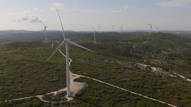 Aerial view of a wind farm with multiple white turbines set amidst rolling green hills and dirt access paths under a clear sky in Portugal.