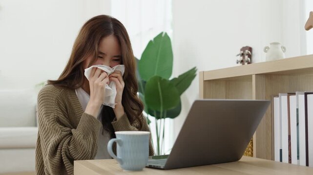 Young woman sneezes into a tissue while working on a laptop at home, illustrating cold or allergy symptoms during remote work.