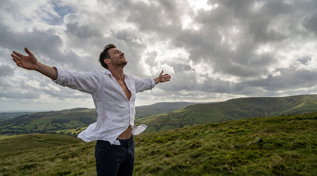 Caucasian man in white linen shirt, standing with outstretched arms and closed eyes, inhaling deeply on a grassy hill, rolling green countryside under cloudy sky, wellness lifestyle photography
