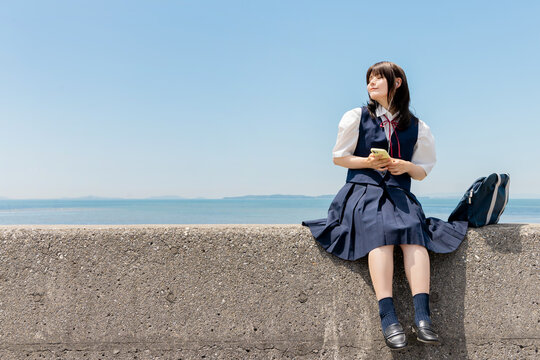 海辺の堤防に座りスマートフォンで音楽を楽しむ女子高校生