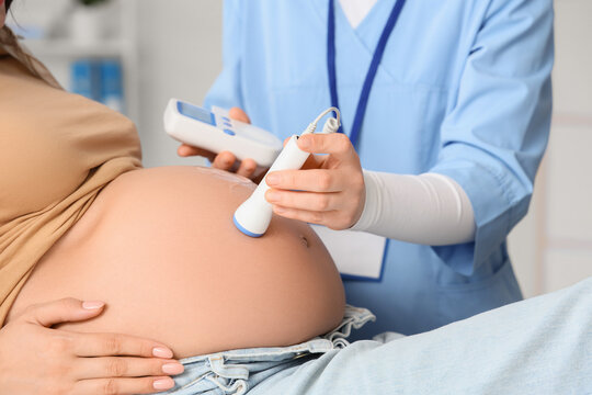 Young pregnant woman and doctor with fetal doppler listening to babys heartbeat in clinic, closeup