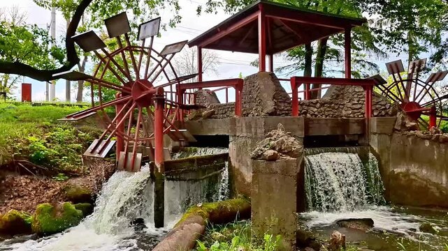 A charming water wheel mill beside a cascading stream in a peaceful park setting. Surrounded by lush greenery and flowing water, this tranquil nature scene offers timeless rural beauty