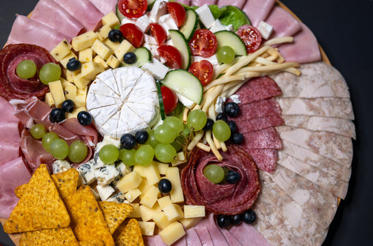 Top view of a charcuterie board with assorted cheeses, ham, salami, head cheese, smoked cheese, cherry tomatoes, cucumber, grapes, tortilla chips and appetizers.