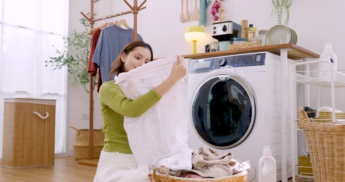 Young Asian woman raises white cloth from basket, moves toward washing machine, prepares laundry cycle, organizing garment care during productive domestic routine in bright cozy apartment room