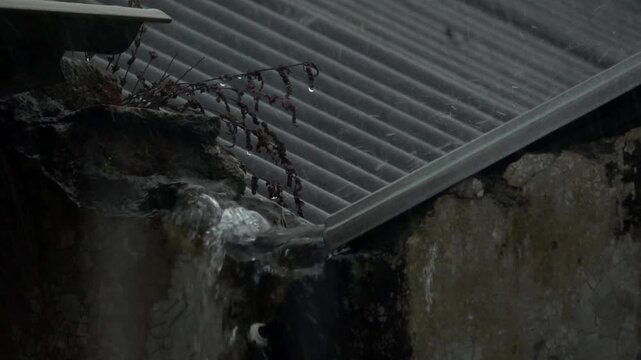 Close up slow motion shot of torrential rainwater gushing and splashing down from a broken corrugated roof corner during a heavy tropical storm.
