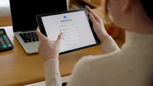 Person checking order tracking on a tablet beside cardboard boxes, managing online shipping and deliveries in a home office.