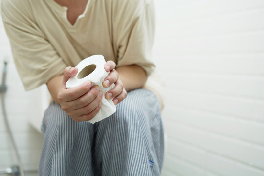 portrait of a man suffers from diarrhea his stomach painful. ache and problem. hand hold tissue paper roll in front of toilet bowl. constipation in bathroom. Hygiene, health care concept.	