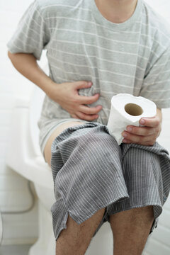 portrait of a man suffers from diarrhea his stomach painful. ache and problem. hand hold tissue paper roll in front of toilet bowl. constipation in bathroom. Hygiene, health care concept.	