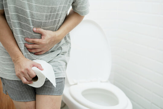 portrait of a man suffers from diarrhea his stomach painful. ache and problem. hand hold tissue paper roll in front of toilet bowl. constipation in bathroom. Hygiene, health care concept.	