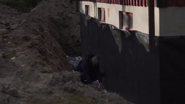 Construction worker checks basement foundation waterproofing and exterior insulation on a house renovation site.