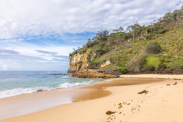 Obraz premium The sandy beach and the southern headland of the large cove with its high, steep sandstone cliffs and coastal vegetation at Steamers Beach in Booderee National Park in New South Wales, Australia.