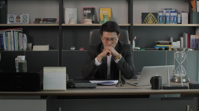 A businessman in a suit sitting at an executive desk with his hands clasped in a serious thinking pose in a modern office at night.